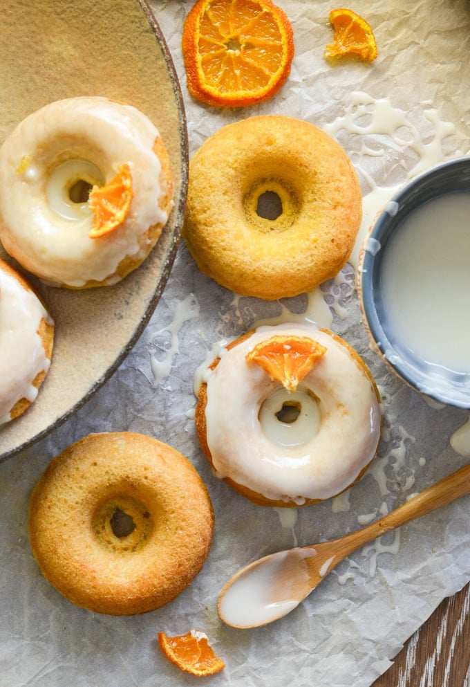 orange olive oil baked donuts on baking parchment with a bowl of frosting and a spoon