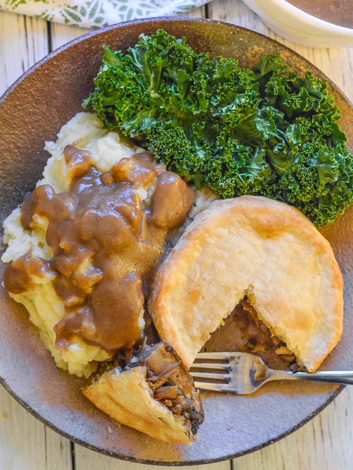 Easy Vegan Mushroom Pie on a plate with a slice taken out. Sides of mashed potato and kale. Gravy too.