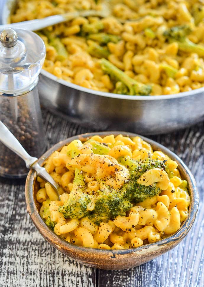 Dairy free mac and cheese with broccoli and black pepper in a small brown bowl, next to a pepper pot and with the pan full of mac and cheese in the background