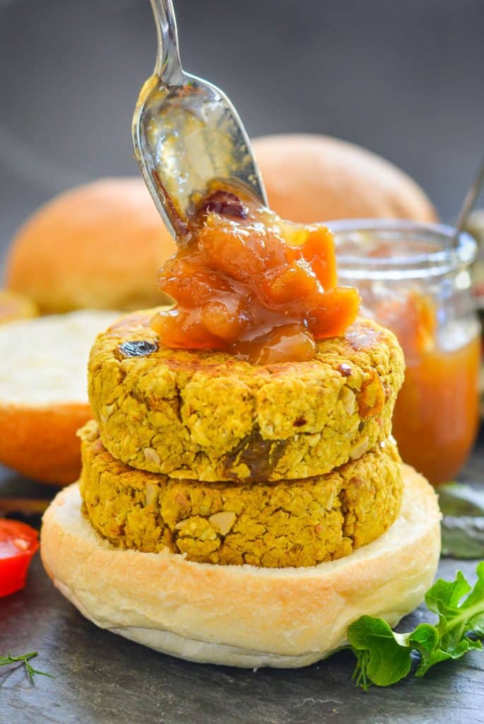 two curried chickpea burgers stacked on a bun with a generous spoon of mango chutney being dolloped on top