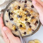 Hands holding a bowl of Overnight Oats topped with banana, nuts and chocolate