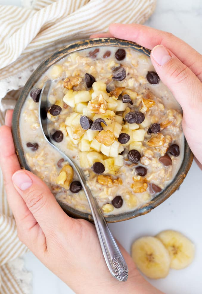Hands holding a bowl of Overnight Oats topped with banana, nuts and chocolate