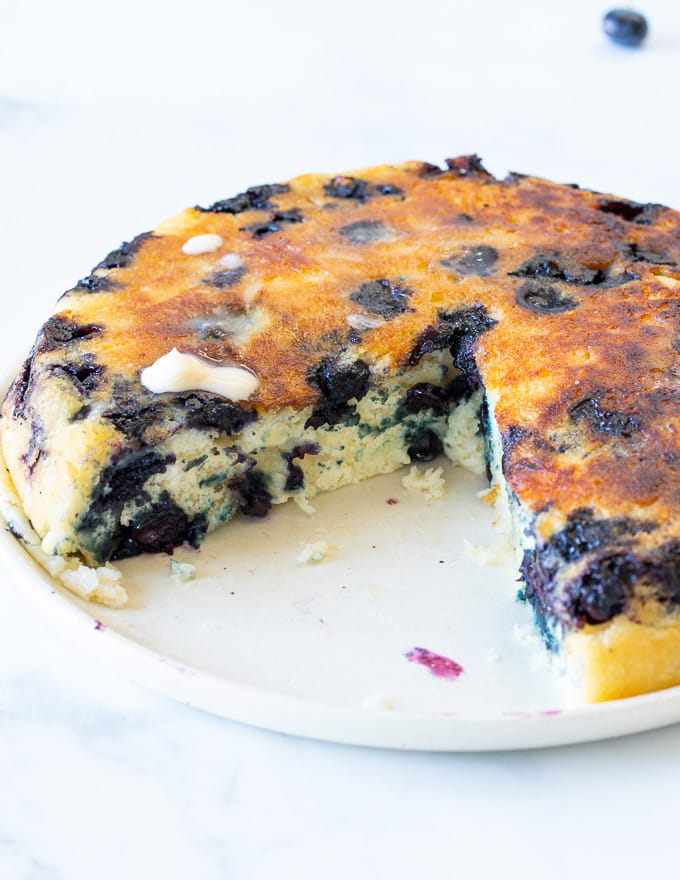 a giant blueberry vegan pancake on a plate with a slice taken out and fluffy inside showing