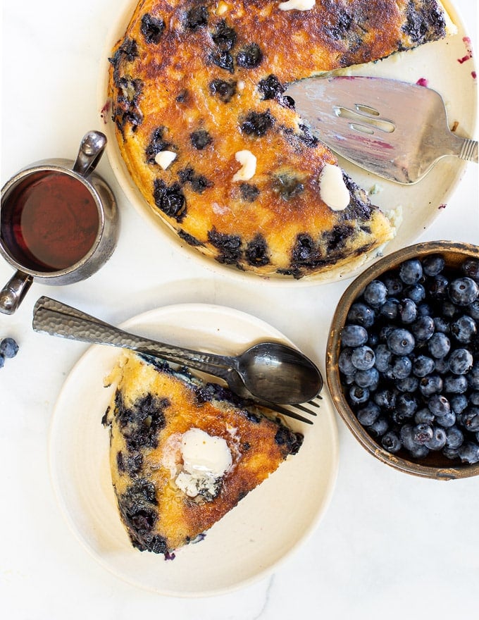 overhead photo of a giant blueberry vegan pancake with a slice on a plate, a bowl of blueberries and a jug of maple syrup