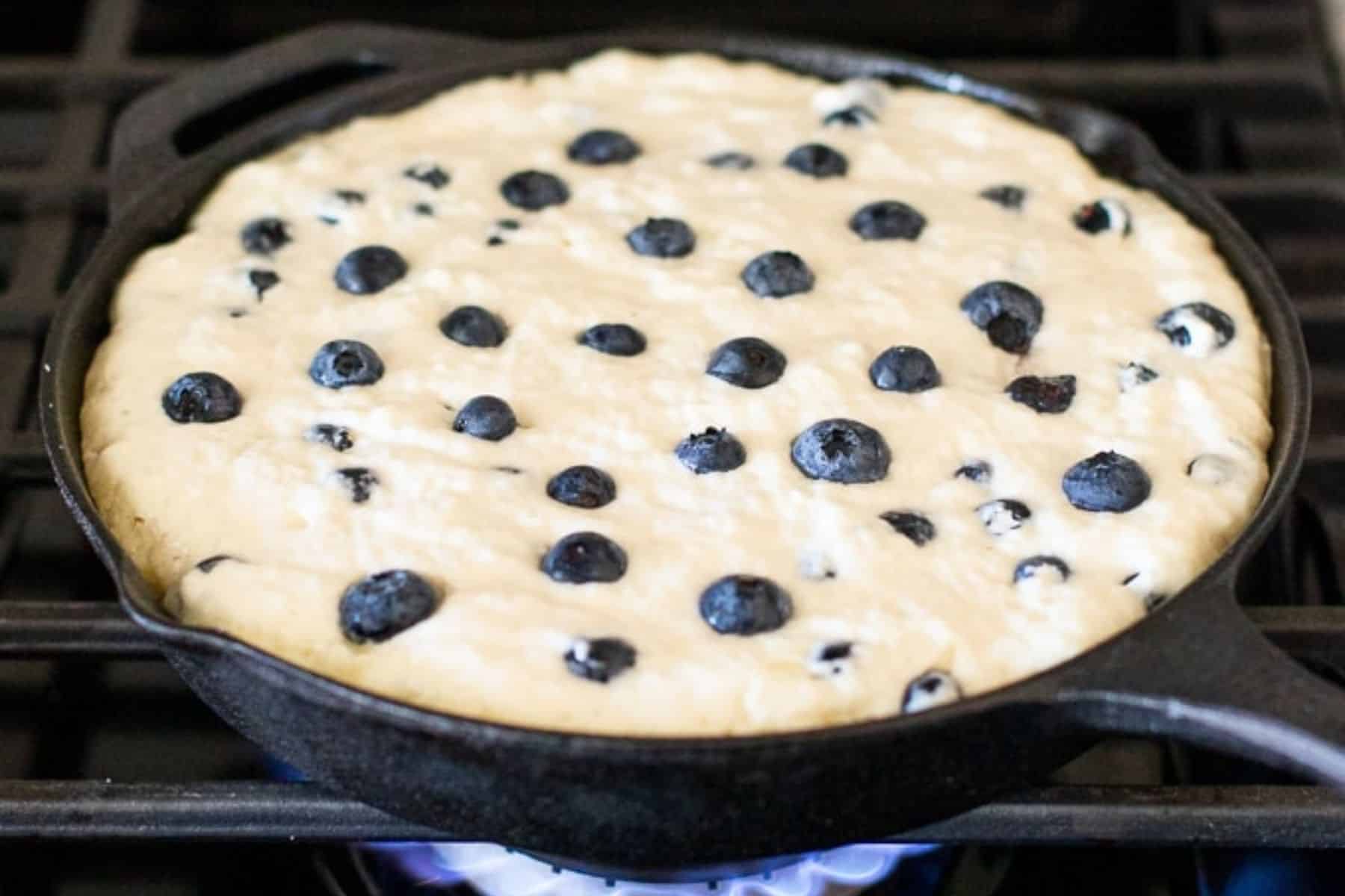 a partially cooked giant vegan blueberry pancake in a cast iron skillet.