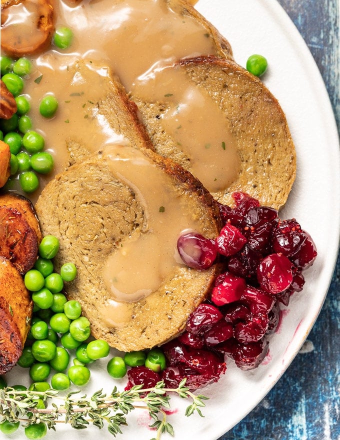 close up picture of vegan roast sliced on a plate with gravy, cranberry sauce and peas