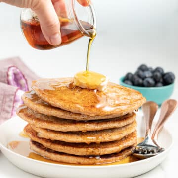 Maple syrup being poured from a small glass jar over a stack of vegan spelt pancakes