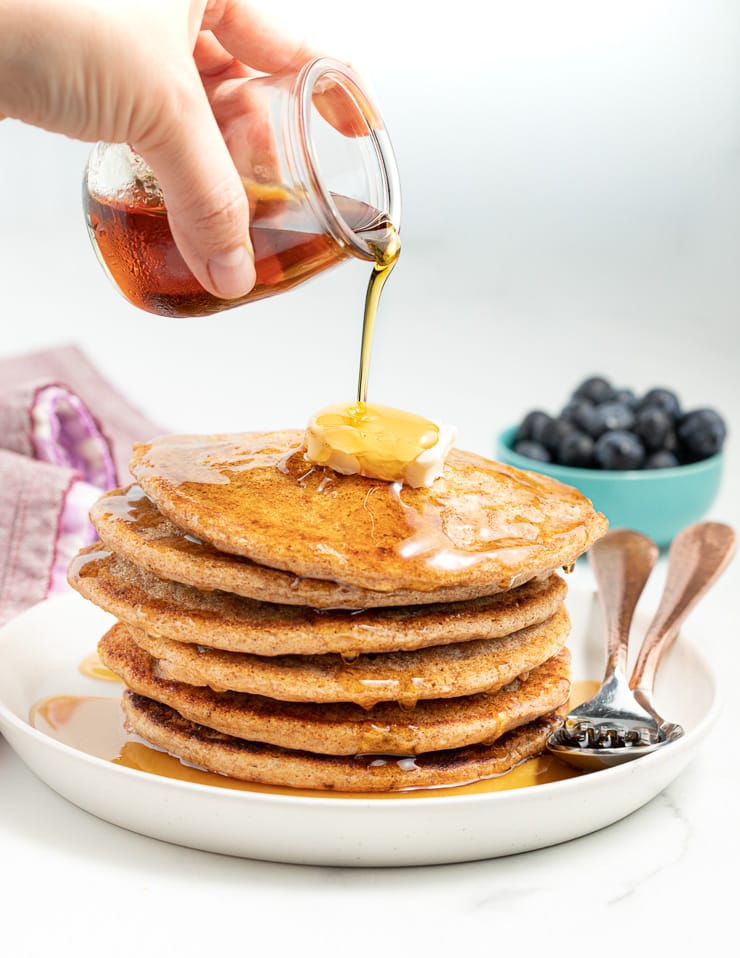 Maple syrup being poured from a small glass jar over a stack of vegan spelt pancakes
