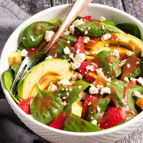 a bowl of strawberry spinach salad with avocado and vegan feta drizzled in strawberry vinaigrette