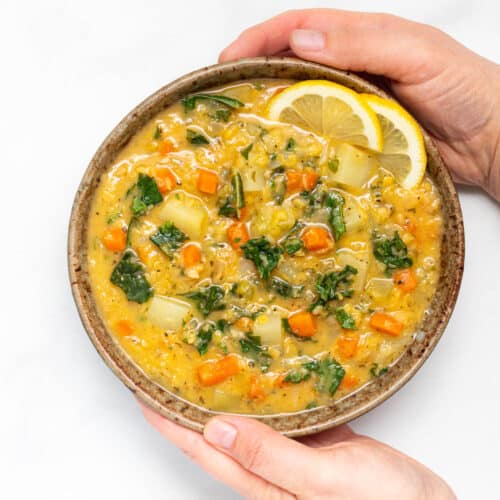 A bowl of lemony red lentil soup with a lemon slice garnish being held by 2 hands