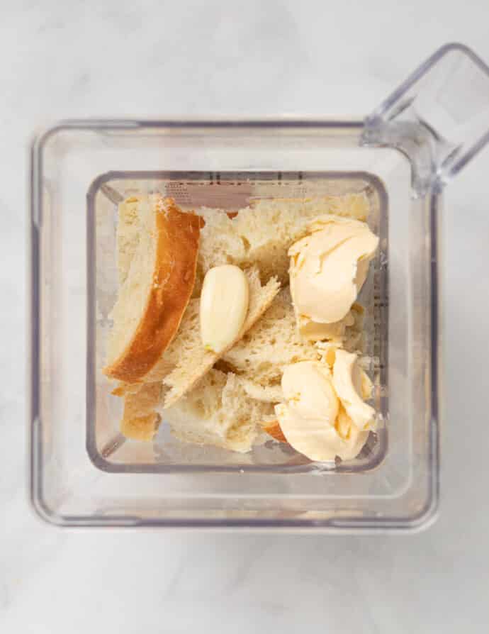 a blender jar with broken up bread, vegan butter and garlic