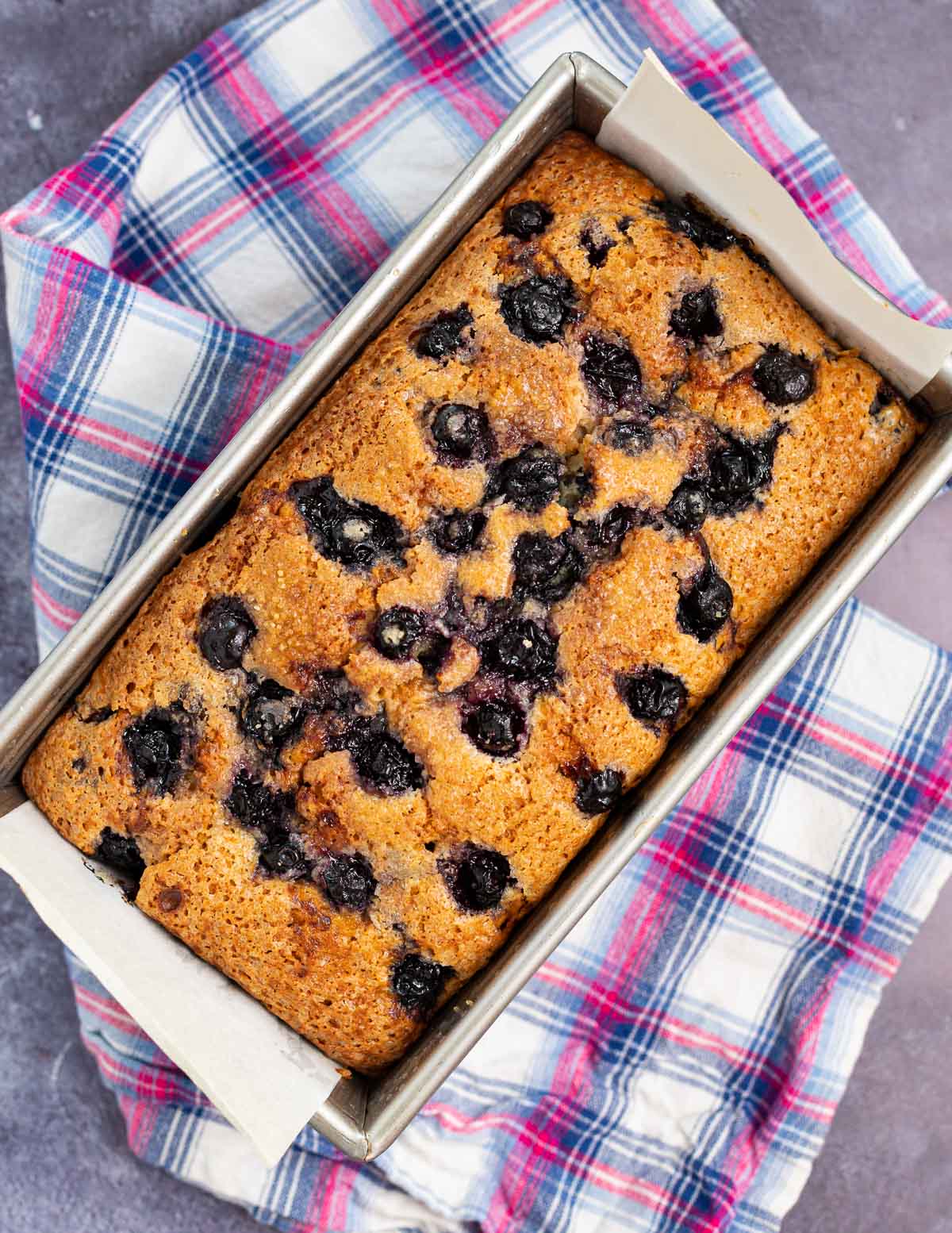 a golden blueberry cake in a loaf pan on a checked cloth