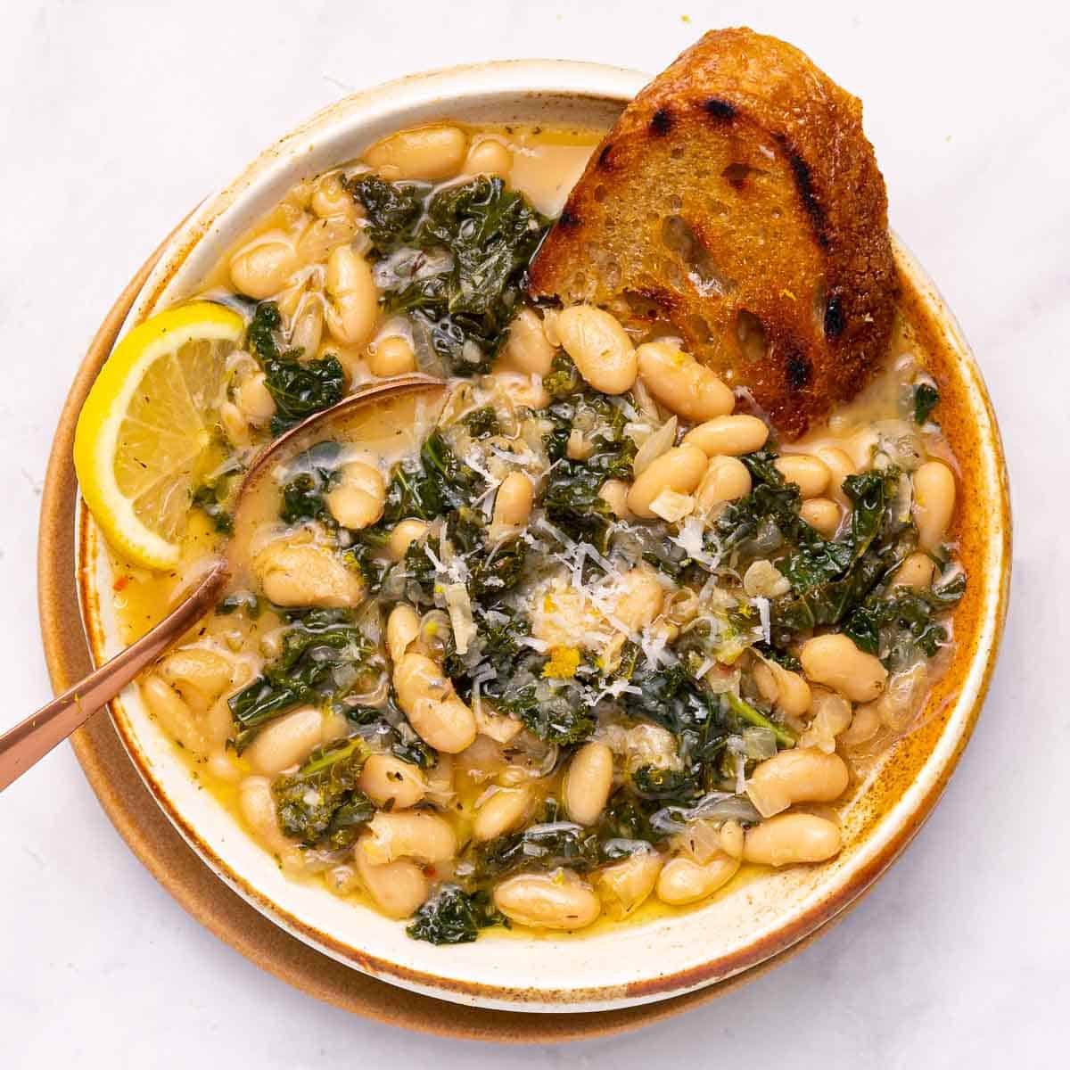 a bowl of braised white beans & greens with a slice of lemon and a piece of toast.