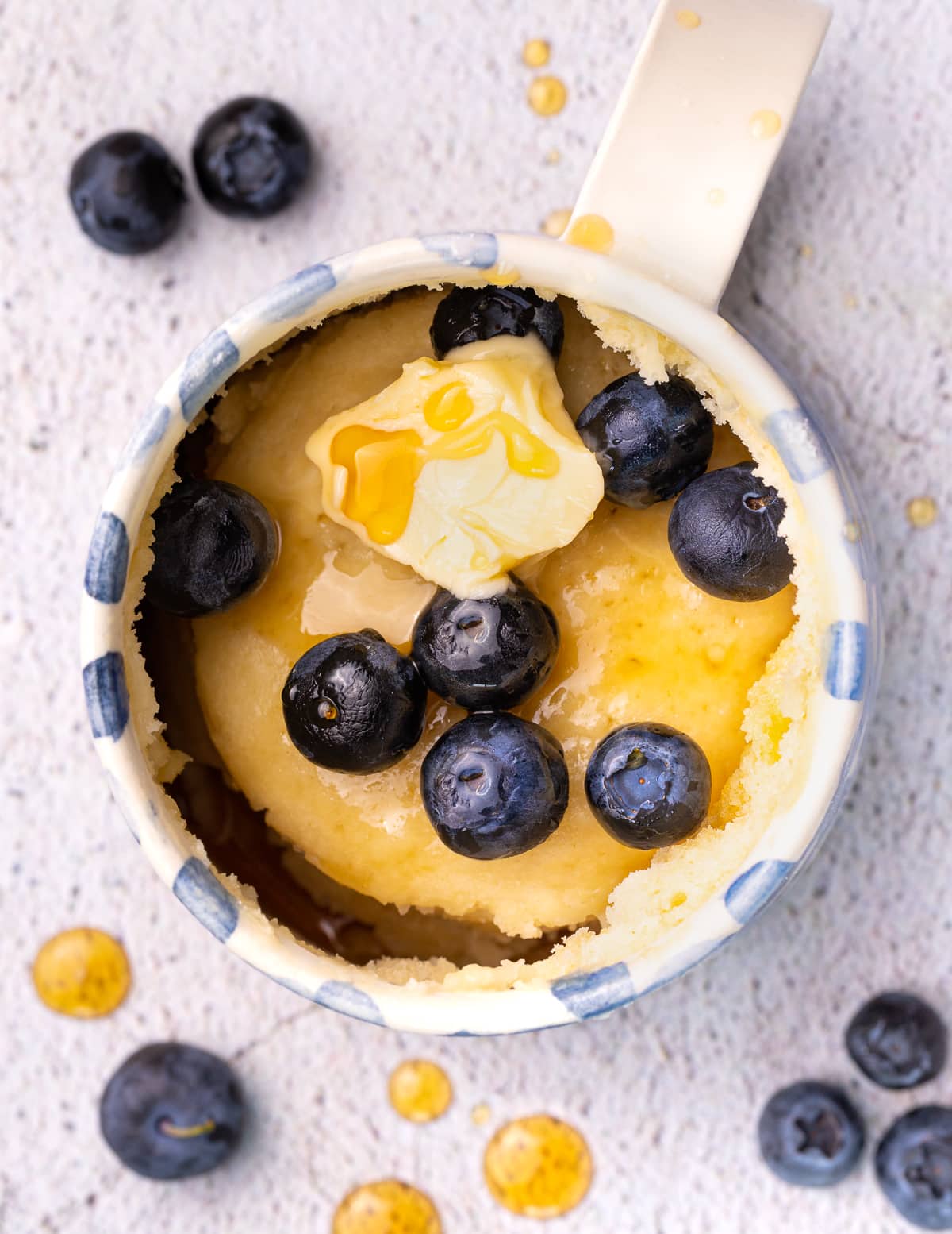 a pancake in a blue and white mug, topped with blueberries, butter an syrup.