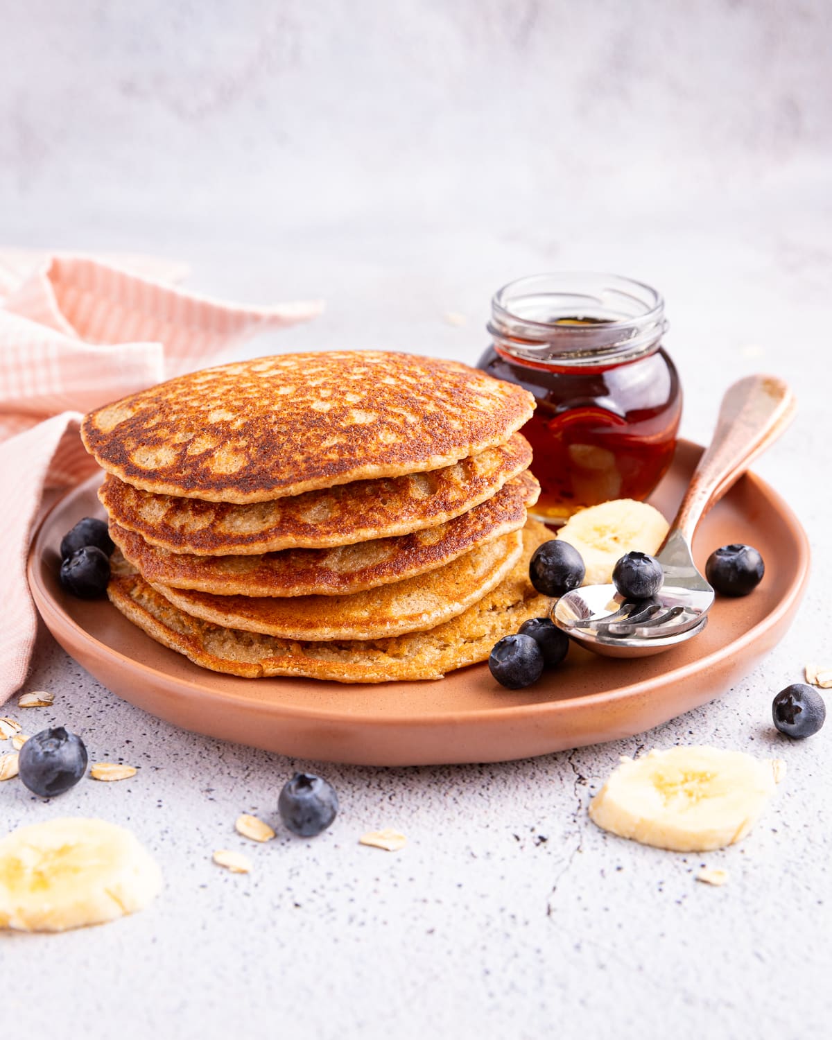 a stack of banana oatmeal pancakes on a plate with a jar of maple syrup, some banana slices, and blueberries. 