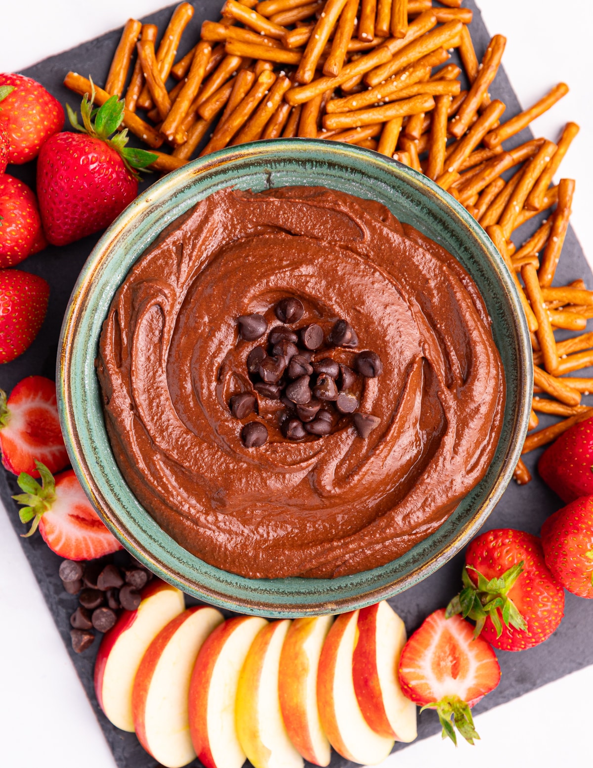 a bowl of dark chocolate hummus surrounded with pretzels, strawberries, an sliced apple