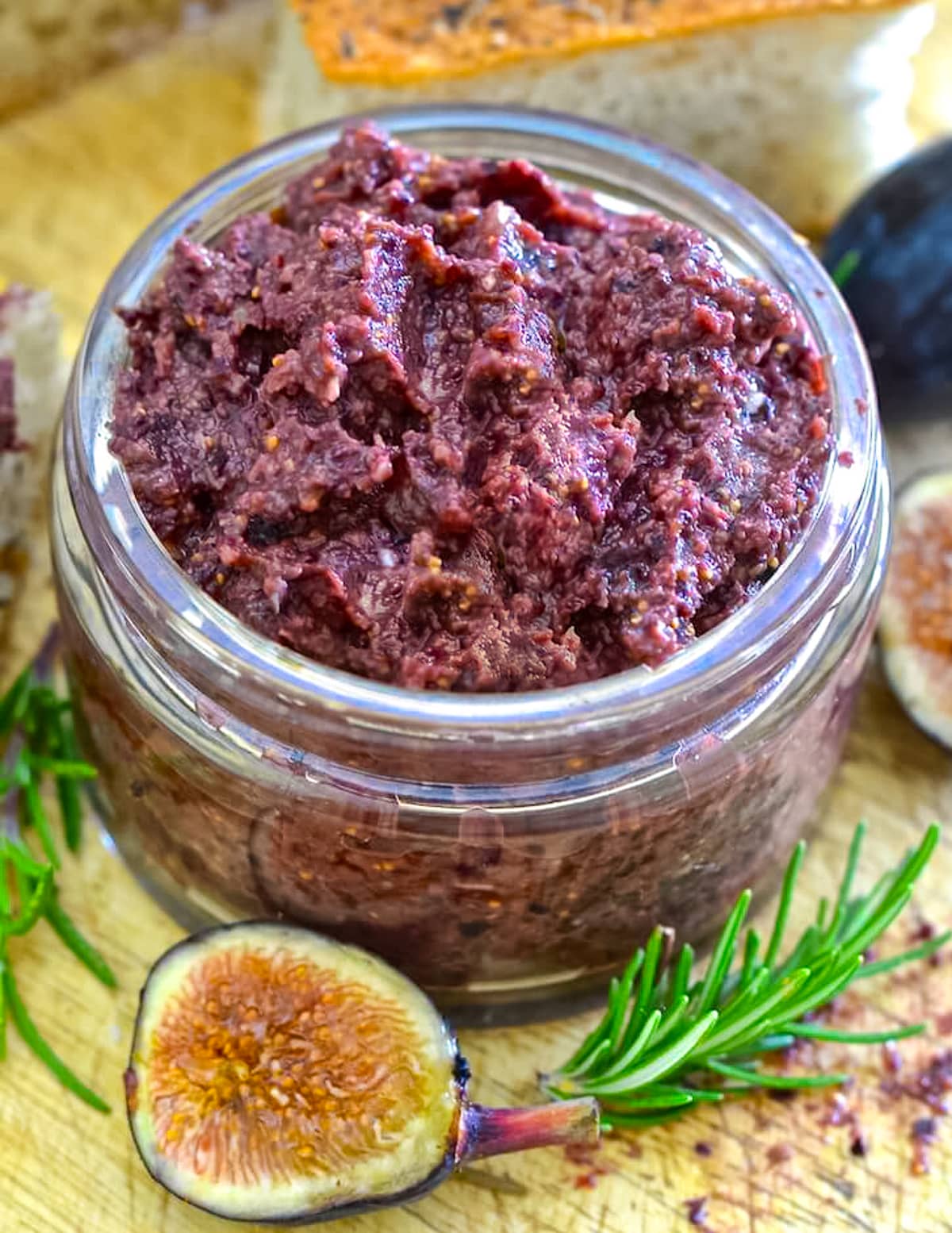 a jar of fig & black olive tapenade with half a fig and rosemary sprig on a wooden board