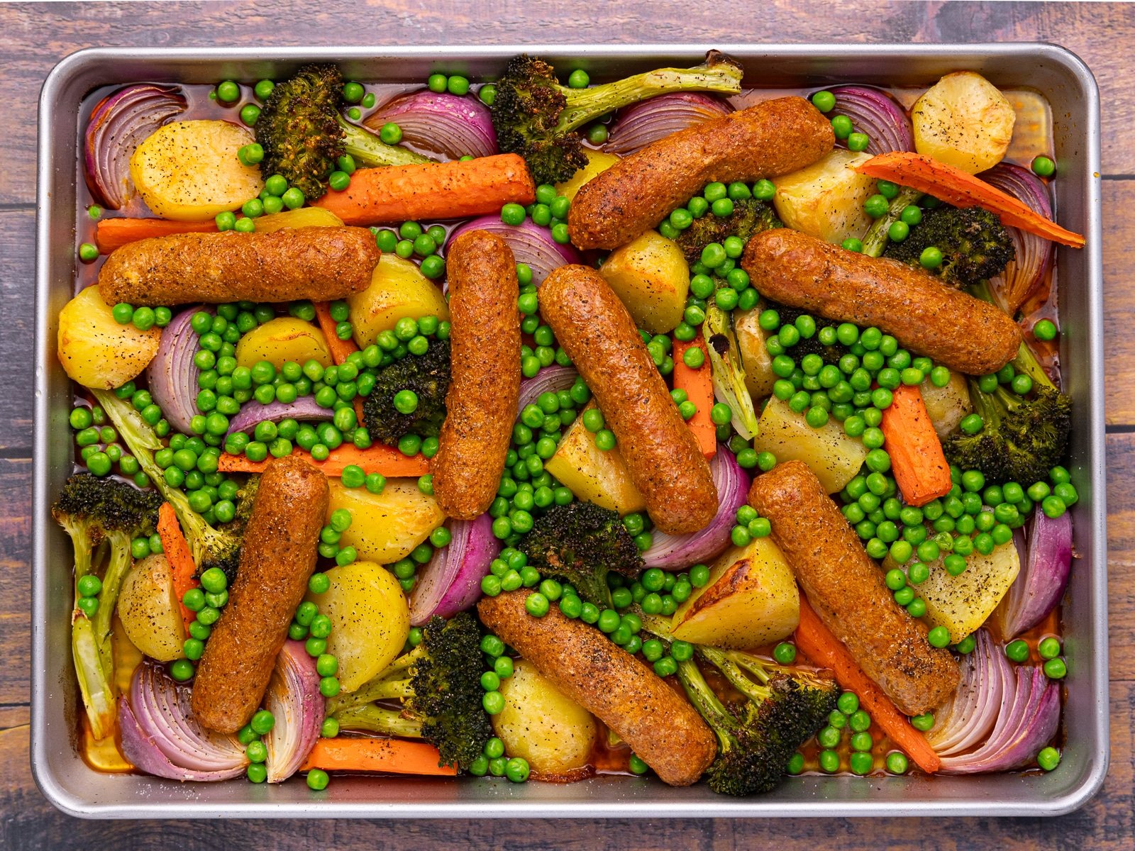 an almost cooked sheet pan meal with sausages, potatoes, broccoli, carrots, red onion, and peas