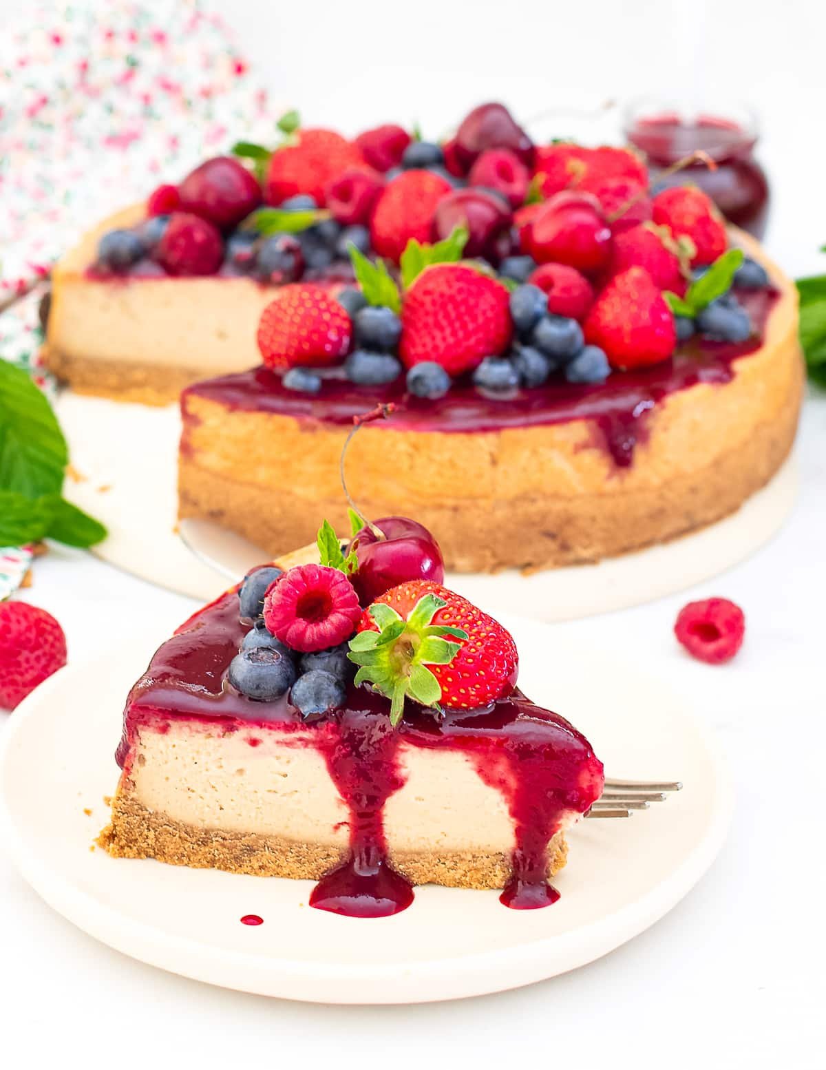 a slice of cheesecake with berry compote and fresh berries on a plate and the remaining large cheesecake tin the background.