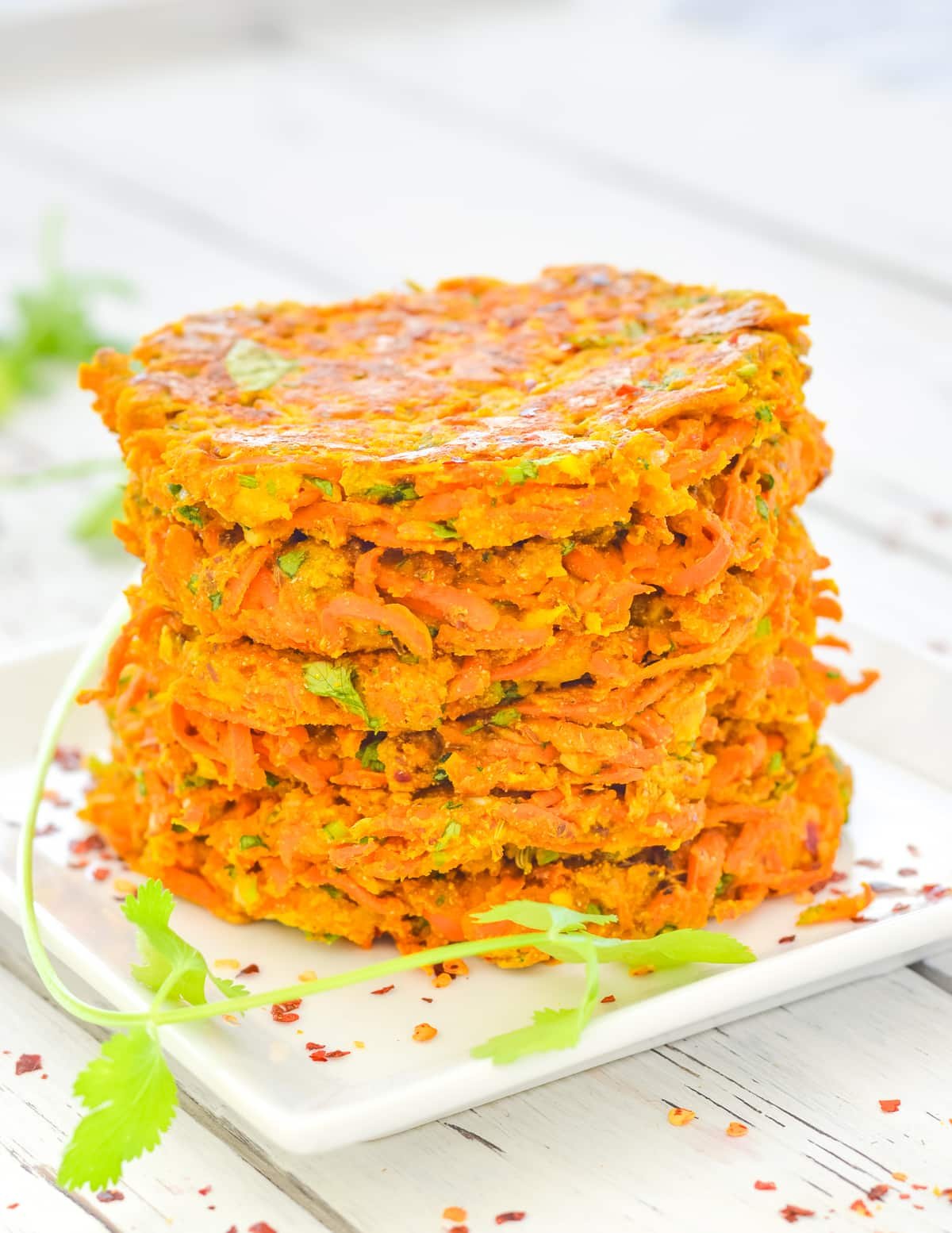 a stack of carrot fritters on a square white plate with a stalk of cilantro for garnish. 