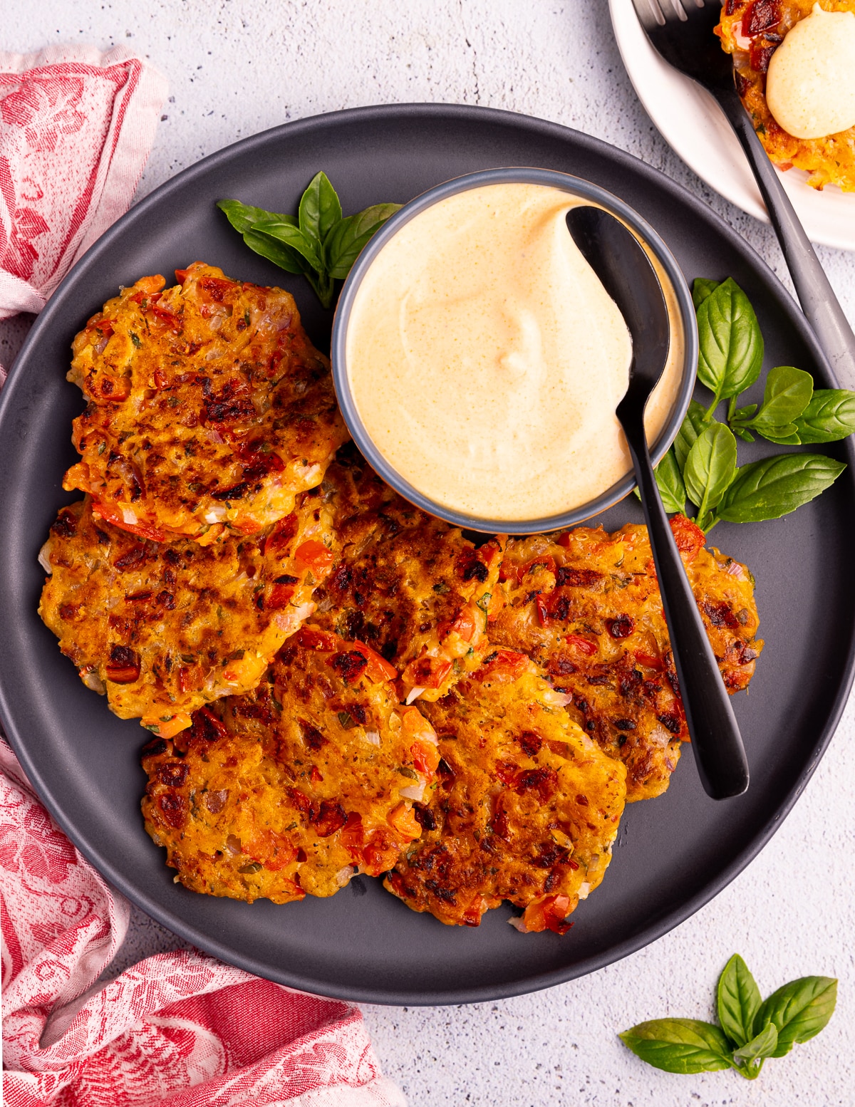 a black plate with a bowl of creamt sriracha dip and lots of golden tomato fritters. Garnished with some fresh basil. 