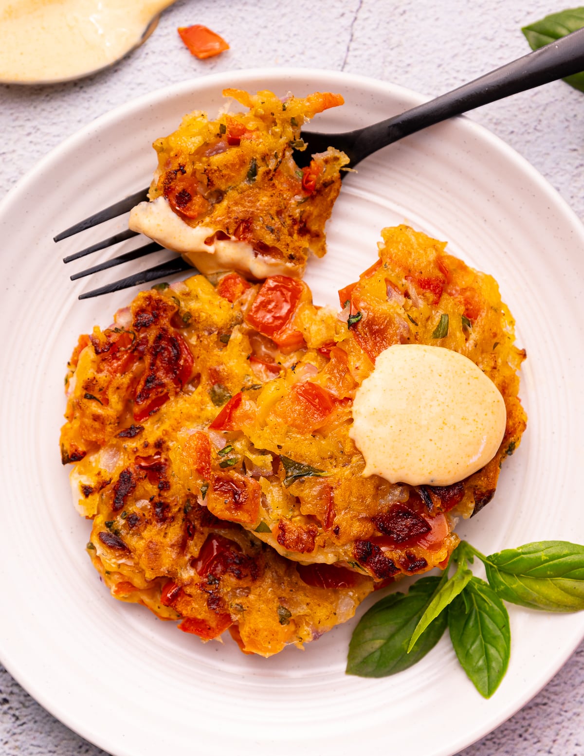 2 tomato fritters on a white plate with a dollop of  creamy sriracha sauce, a black fork, and a sprig of fresh basil. 