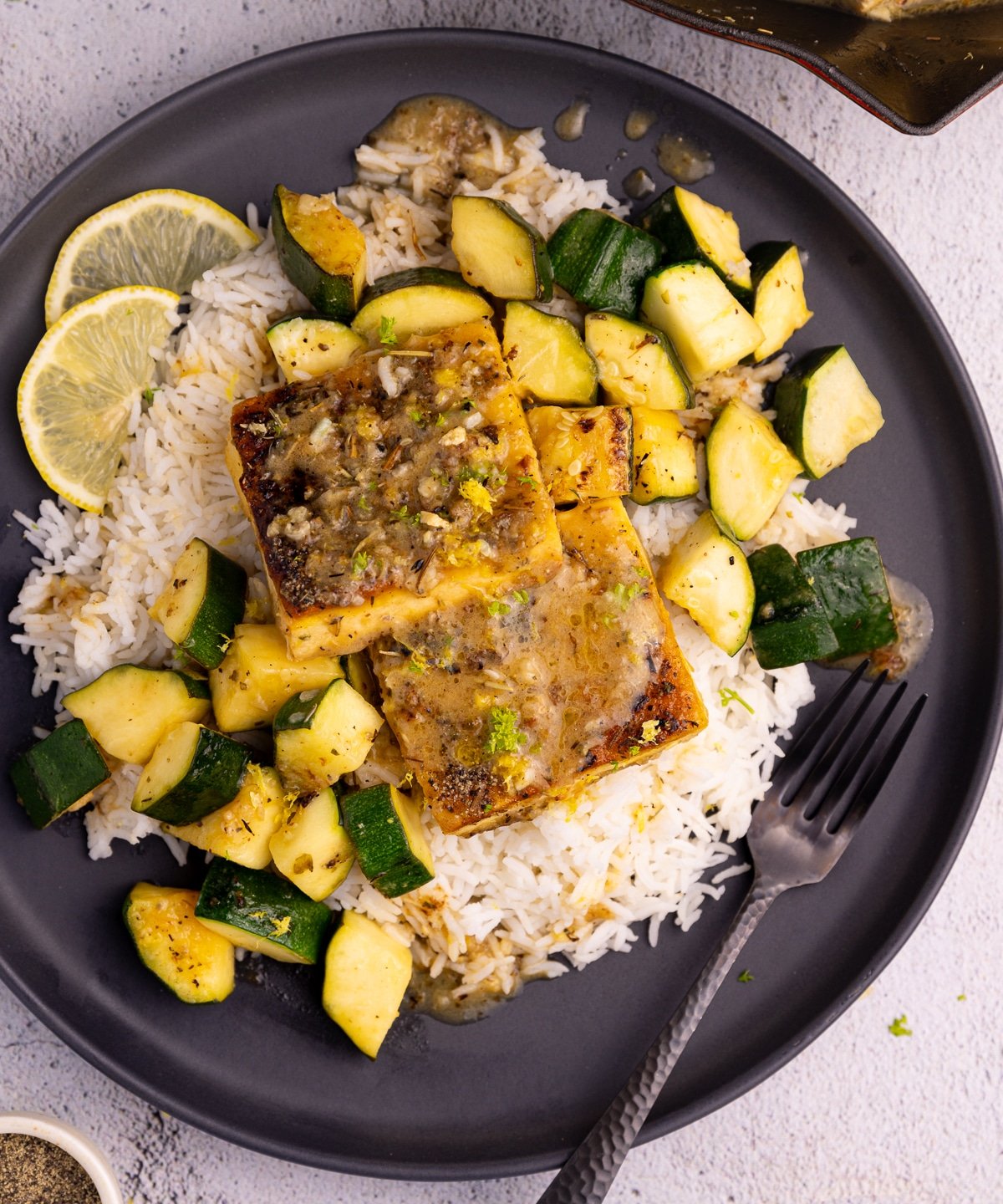 A black plate piled up with rice, golden, crusty, herby tofu squares, and cubed zucchini, sauce and a lemon slice garnish. 