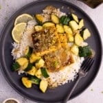 A black plate piled up with rice, golden, crusty, herby tofu squares, and cubed zucchini, sauce and a lemon slice garnish.