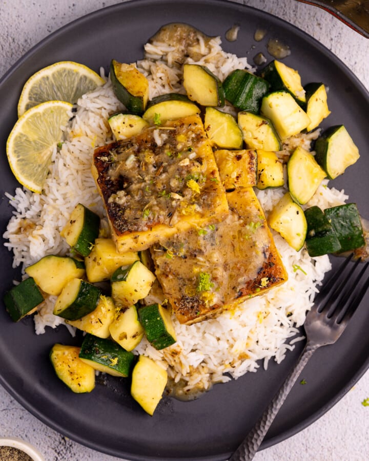 A black plate piled up with rice, golden, crusty, herby tofu squares, and cubed zucchini, sauce and a lemon slice garnish.