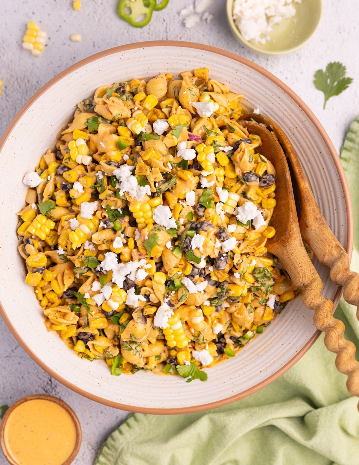 a large salad bowl full of Vegan Elote Pasta Salad, scattered with vegan feta cheese, with some wooden salad servers tucked in the side of the bowl. 