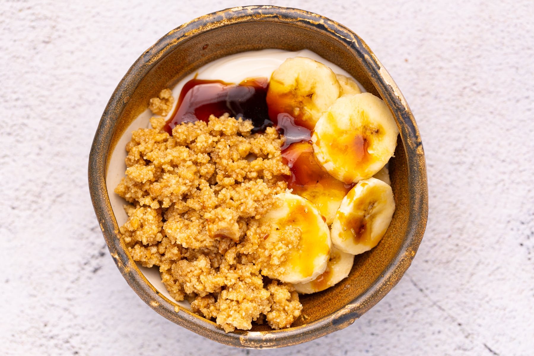 A brown bowl filled with yogurt, sliced banana, buttery cookie crumbles, and drizzles of vegan honey. 