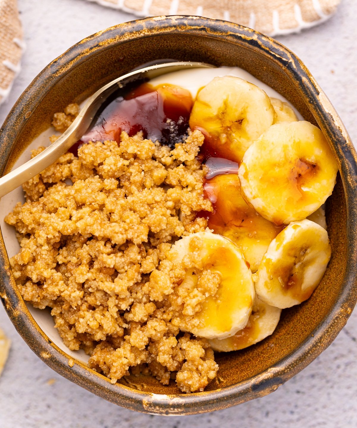 a rustic brown bowl filled with yogurt, buttery cookie crumbles, sliced banana, and vegan honey. 