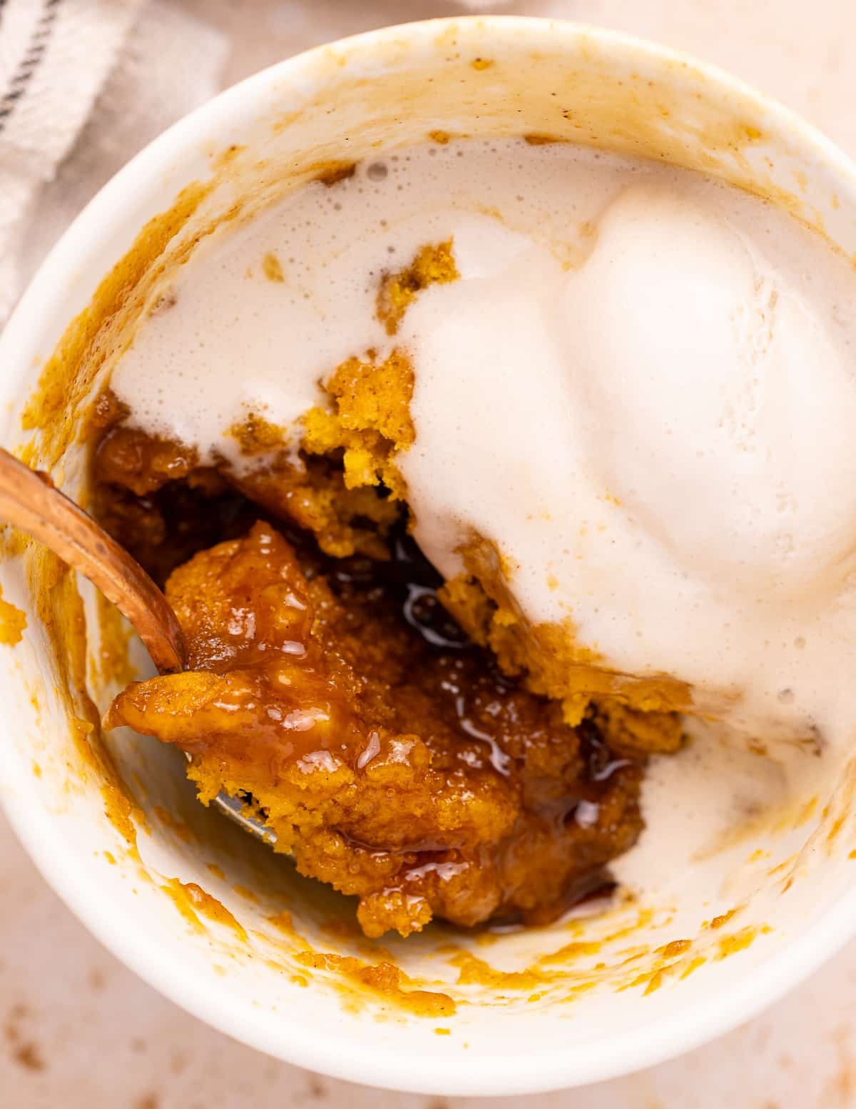 A close up shot of a pumpkin mug cake topped with vanilla ice cream. It has a spoon scooping out a bite and shows the caramel and the fluffy sponge. Yum!