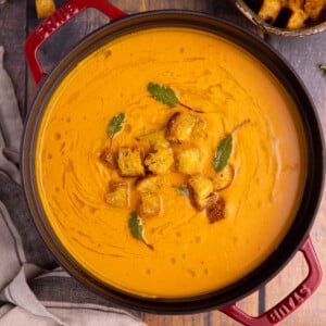 a red pan full of sweet potato soup. Garnished with a drizzle of olive oil, croutons, and fresh sage leaves.