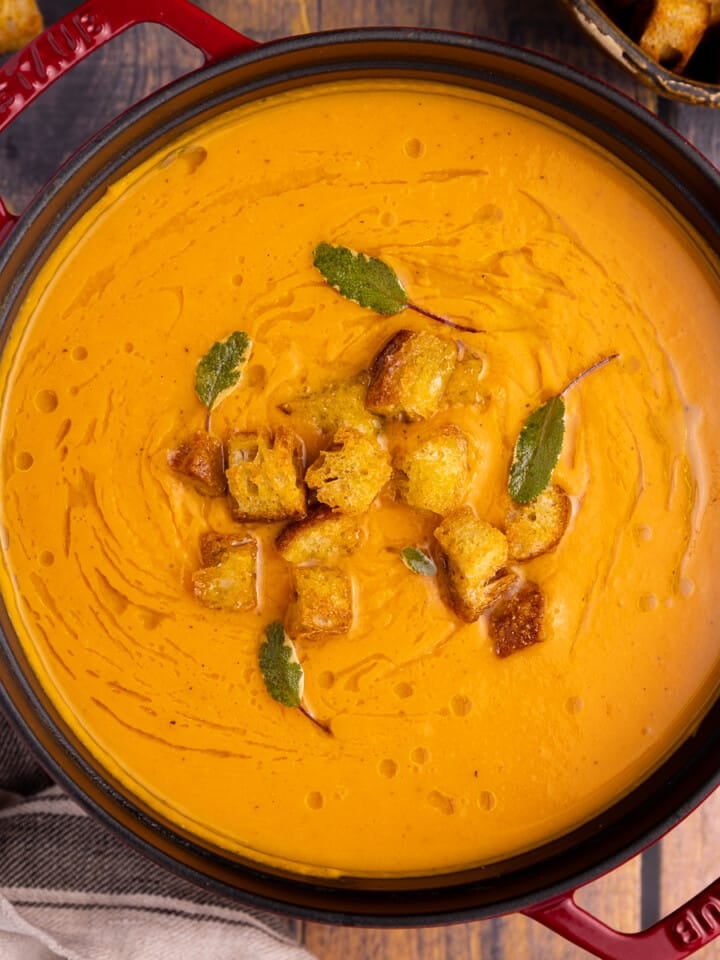 a red pan full of sweet potato soup. Garnished with a drizzle of olive oil, croutons, and fresh sage leaves.