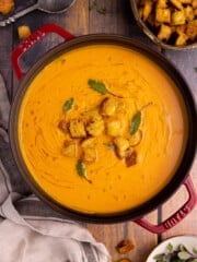 a pan of vegan sweet potato soup that is garnished with croutons, olive oil, and frehs sage leaves.