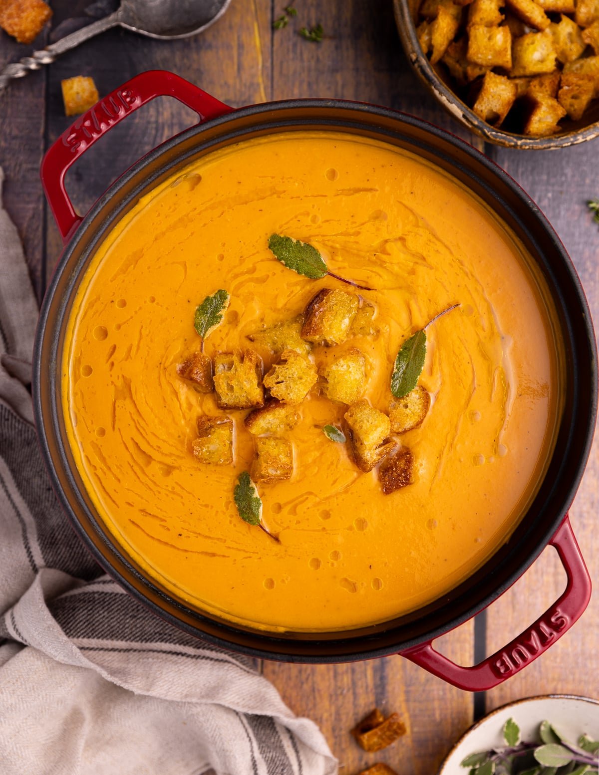 A red Staub pan filled with vegan sweet Potato. soup. The soup is drizzled with olive oil and sprinkled with croutons and fresh sage leaves. 