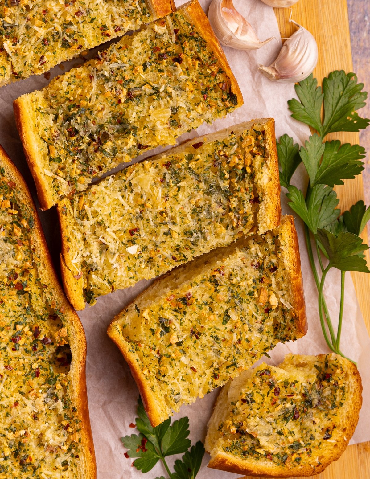 slices of golden garlic bread on a cutting board with fresh garlic cloves and parsley. 